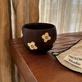 Handmade ceramic mug with gold flower accents, rustic dark glaze, on wooden table near studio pouch
