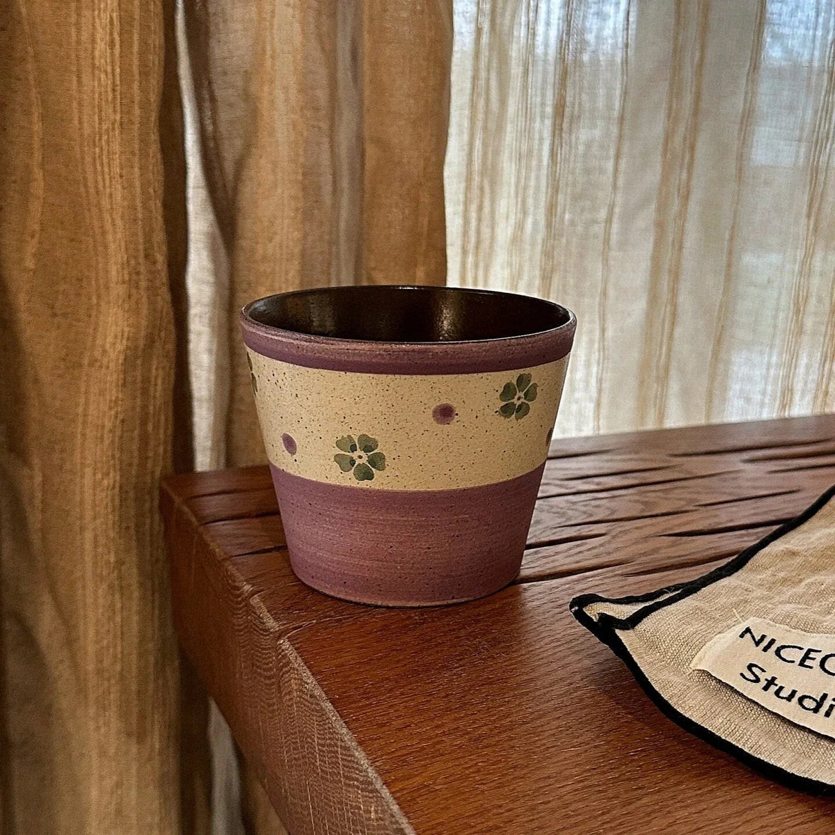 Handmade ceramic mug with green floral pattern, purple rim, and speckled beige band, on wooden table near curtain