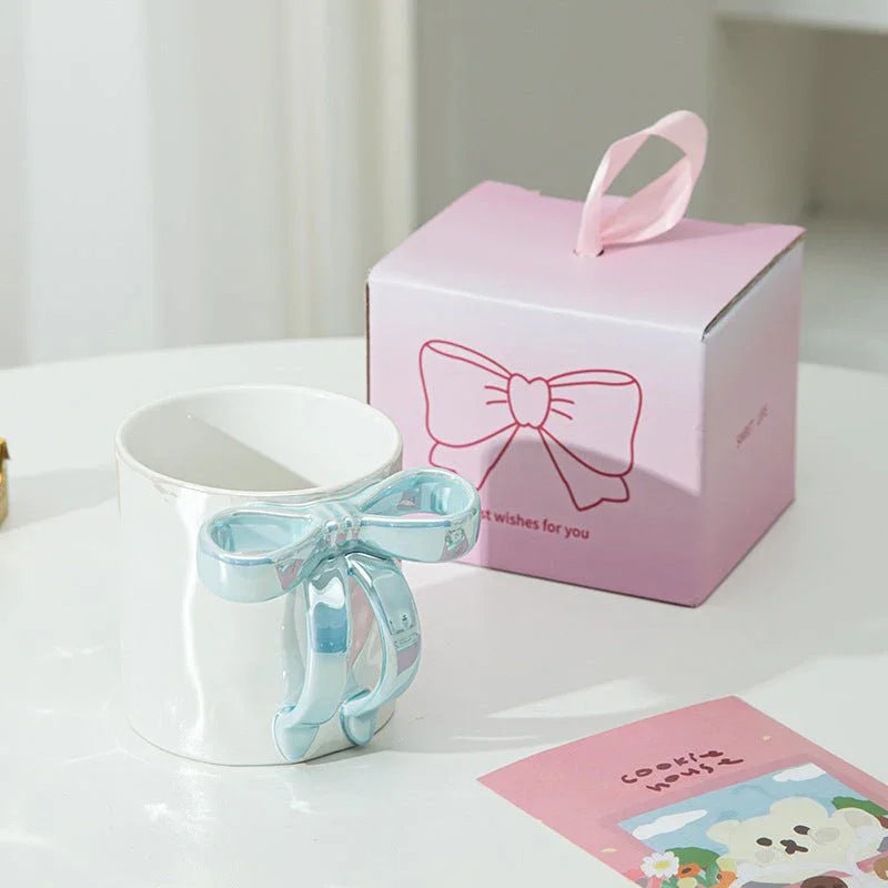 White ceramic mug with shiny silver bow handle beside pink gift box, perfect for tea or coffee