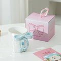 White ceramic mug with blue bow handle beside pink gift box, perfect for tea or coffee lovers